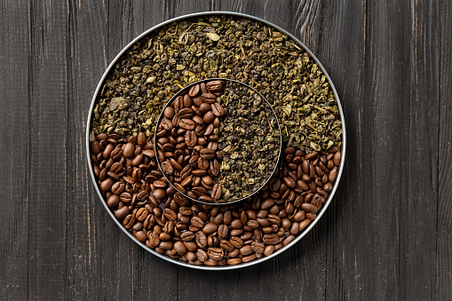 Green dry tea and roasted coffee beans in two round plates of different sizes one on top of the other on a dark wooden background top view close-up. Conceptual composition of green tea and coffee beans on a dark background with space for text.