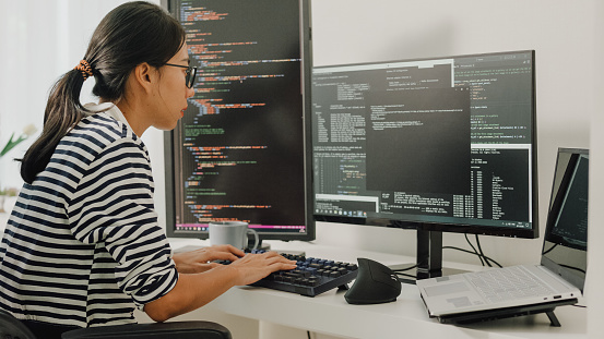Young Asian woman software developers using computer to write code sitting at desk with multiple screens work remotely at home. Programmer development concept.