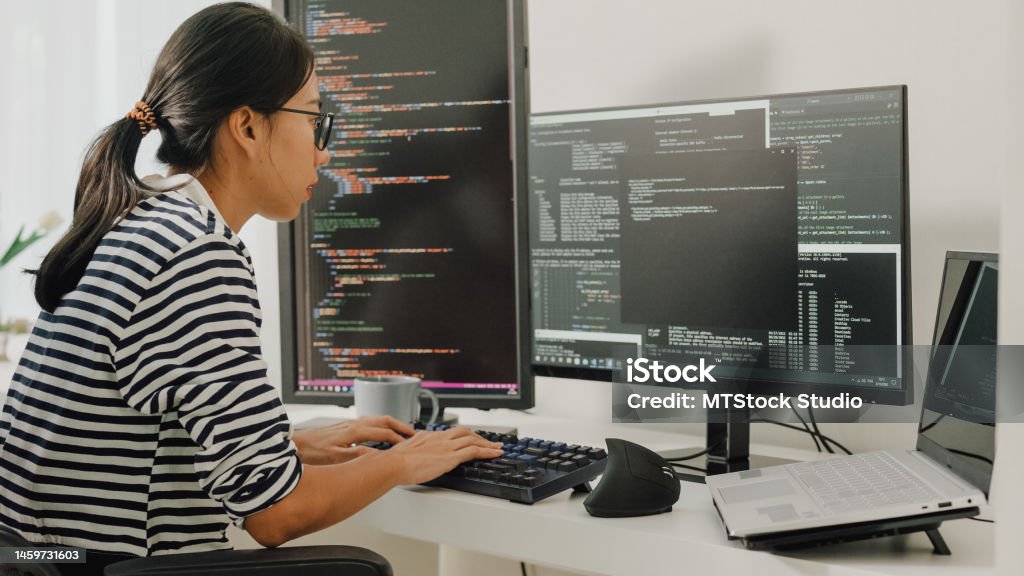 Young Asian woman software developers using computer to write code sitting at desk with multiple screens work remotely at home. Young Asian woman software developers using computer to write code sitting at desk with multiple screens work remotely at home. Programmer development concept. Computer Programmer Stock Photo