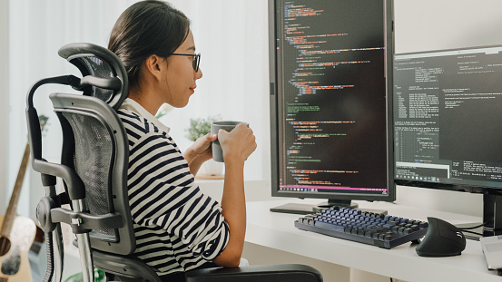 Young Asian woman software developers drinking coffee and using computer to write code sitting at desk with multiple screens work remotely at home. Programmer development concept.