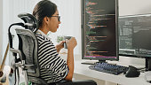 Young Asian woman software developers drinking coffee and using computer to write code sitting at desk with multiple screens work remotely at home.