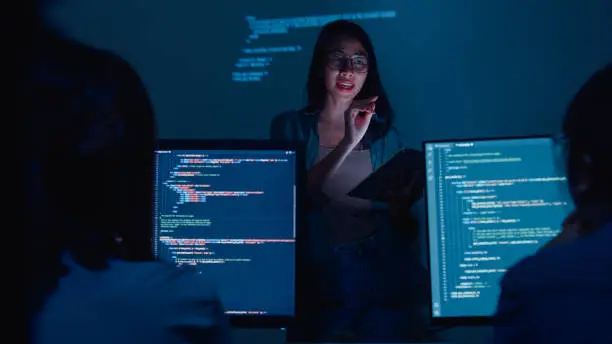 Photo of Young Asian woman software developers mentor leader manager talking to executive team analyzing source code in office at night.