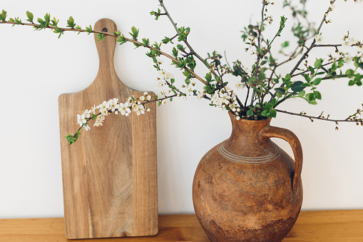 Blooming cherry branches in old vase and wooden board on table against white wall. Spring flowers in kitchen still life. Simple countryside living, home rustic decor. Hello spring