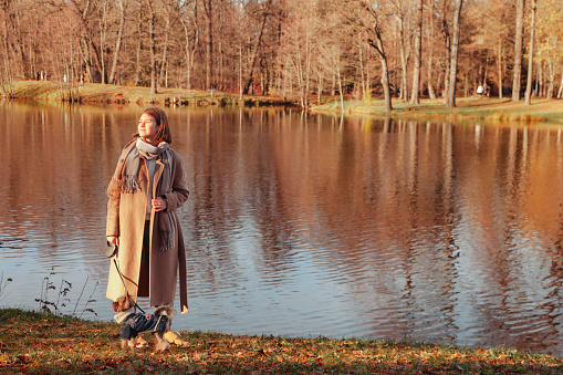 Full length teen girl in beige coat walking with yorkshire terrier dog in autumn park with lake. Pretty young lady with doggy in fall garden outdoor. Pet love concept. Copy text space for advertising