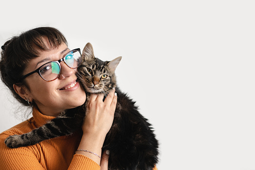 Latin woman wearing eyeglasses is holding her tabby cat while smiling and looking at camera. Isolated white background with copy space for advertisement
