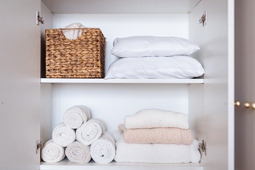 Clothes pillows, towels organized and folded in wicker basket and on shelves in white wardrobe. Space organization and storage system. Tidy up closet