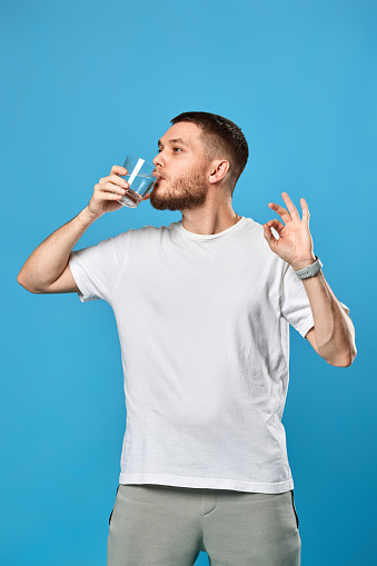 handsome bearded man drinks water from a glass and shows Ok gesture on blue background