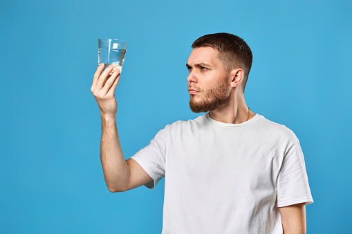 Young guy carefully looks at a glass of water and examines the purity of the water on blue background