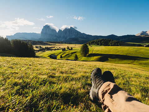 A man is resting admiring the view in Seiser Alm at sunrise - Famous landmark in northern Italy. Italian Dolomites landscape. Pov view with the man's legs.