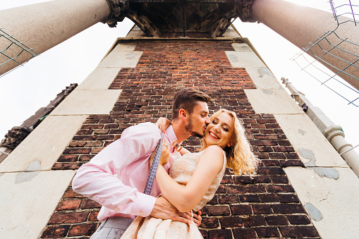 A young boyfriend kisses his girlfriend in a cheek near a brick wall and two columns