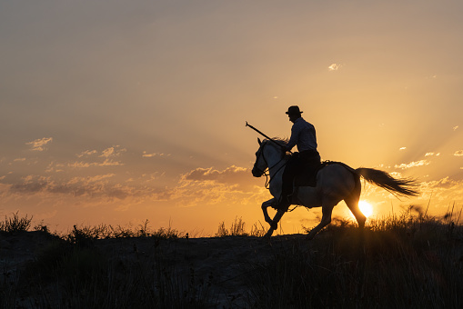 Saintes-Maries-de-la-Mer, Bouches-du-Rhône, Provence-Alpes-Cote d'Azur, France. July 5, 2022. Man with a pike riding a horse at sunrise.
