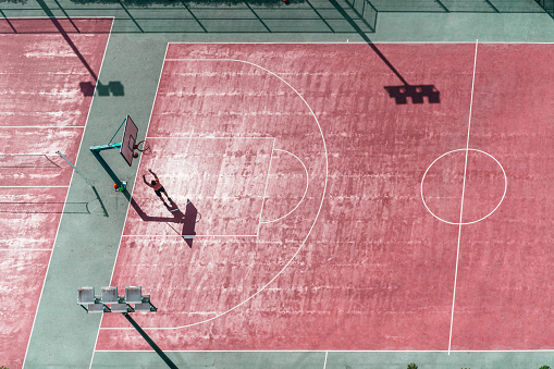 A drone view of a colorful basketball court with a silhouette of a man throwing the ball into the basket; view from the high above of red and green basketball playground field on a sunny day