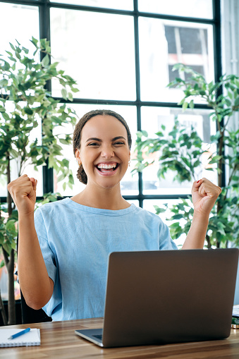 Vertical photo of excited happy mixed race young business woman, sit at a desk with laptop in office, rejoicing in success, big profit, celebrate deal, gesturing with fists, looks at camera, smiling