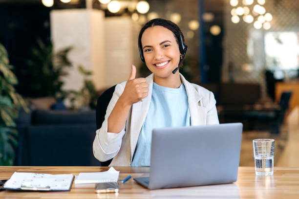 istock Happy successful mixed race woman, with headset, call center agent, support service operator, sits in the office, work distantly, consult clients online, looks at camera, smile, shows thumb-up gesture 1459814679