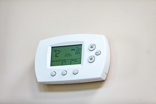Push button automated digital thermostat to control heating and cooling air conditioning temperatures mounted on a residential home living room interior wall.