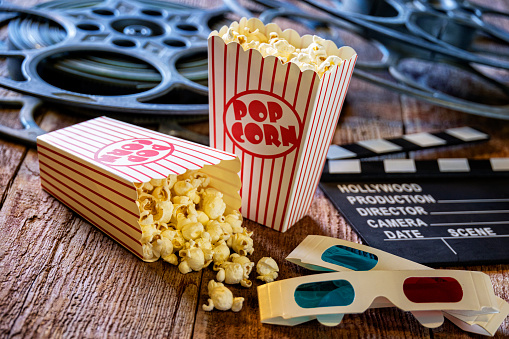 This is a conceptual photo relating to going out to the movies or watching a movie at home. There are two old retro movie reels on a wood background with a movie clap board and two bags of popcorn. There is also a pair of 3-D glasses.