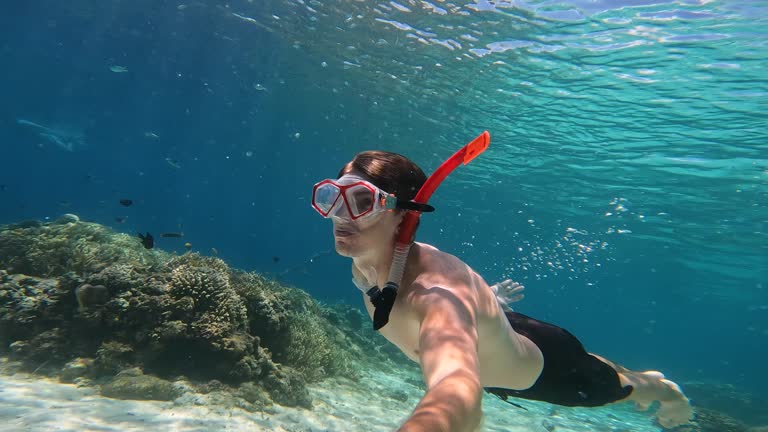 A man in a red snorkel mask dives underwater, swimming next to the white sand at the bottom of the sea. A man on vacation in the Maldives dives under warm sea water. Exploring seabed by amateur diver.