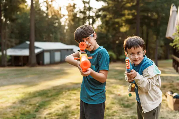 A Eurasian tween boy and his younger brother smile and take aim while having a playful nerf gun battle in the back yard of their home on a warm summer evening. Both boys are wearing protective eyewear and aiming their toy guns directly at the camera.