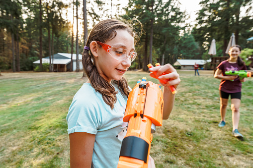 A tween girl smiles and puts foam ammunition in a toy gun while having a playful nerf battle with her friend in the back yard of her rural home on a warm evening.