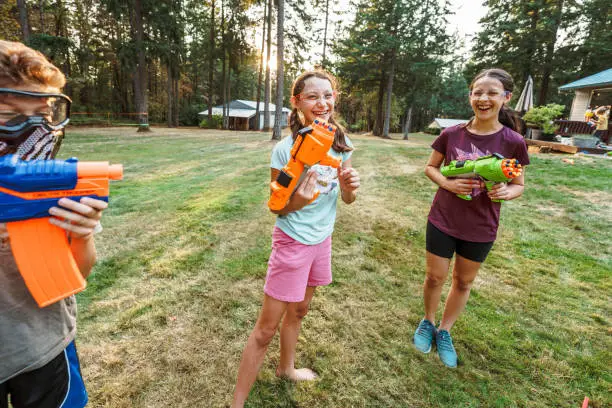 A multiracial group of boys and girls stand outside in the grass and laugh while having a playful battle with toy nerf guns. The kids are wearing protective eyewear.
