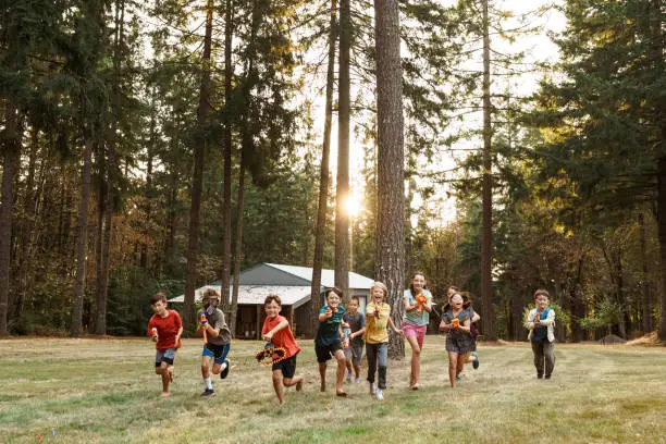 Wide shot of an excited group of kids running in a large rural back yard toward the camera while having a playful nerf gun battle.