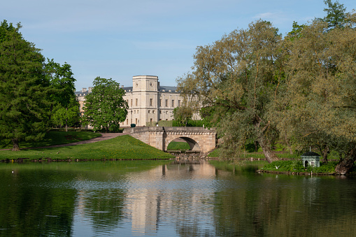 View of the Karpin Bridge over the cascade between the Karpin Pond and the White Lake of Gatchina Park and the Gatchina Palace in the background on a sunny summer day, Gatchina, St. Petersburg Russia