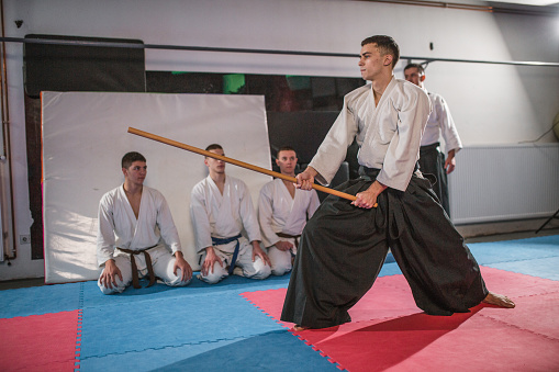 A young aikido martial artist holds a wooden sword in a gym and demonstrates aikido techniques to a group of fighters