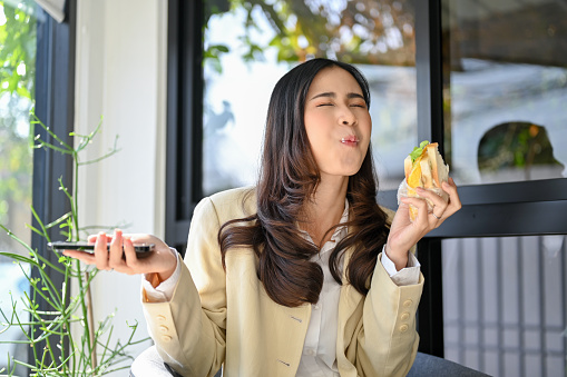 Joyful, happy and hungry millennial Asian businesswoman enjoying with her healthy breakfast, eating a sandwich at the cafe. happiness overload from yummy food