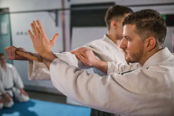 A young aikido martial artist holds a wooden sword in a gym and demonstrates aikido techniques to a group of fighters