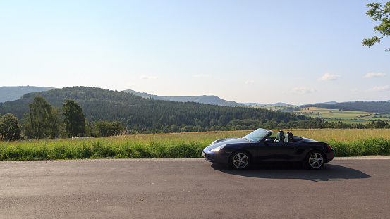 Gersfeld, Germany - July 23, 2021: Blue roadster Porsche Boxster 986 with Wasserkuppe panorama in Rhön Mountains. The car is a mid-engine two-seater sports car manufactured by Porsche.