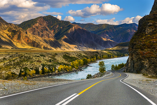 Serpentine asphalt road among high snow-capped mountain peaks, yellow desert, autumn green forest and blue sky. Car on the highway against the background of a mount landscape.