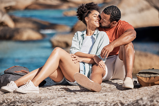 Love, kiss and happy couple on a rock at the beach relaxing, bonding and embracing while on vacation. Happiness, smile and young man and woman by the ocean on a romantic, calm and zen seaside holiday