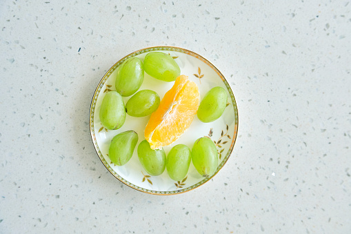 Fresh grains of white grapes and a petal of orange are placed in a golden patterned plate and they are placed on a white marble table.