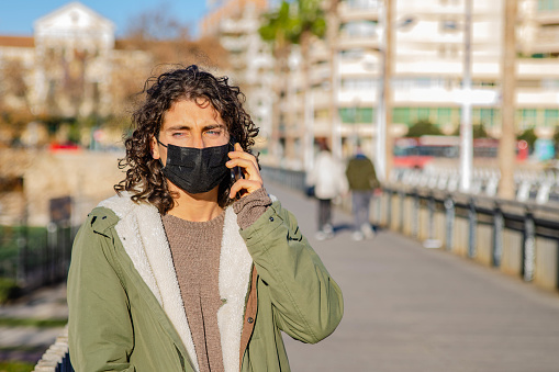 Picture of Young Curly Long Hair Man With Mask and Green Jacket Talking to the Phone with Disgusted Face Expression Hearing Bad News at the Bridge.