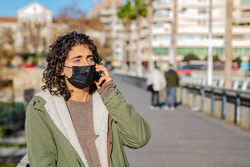 Picture of Young Lifestyle Man With Worried Expression Face Wearing Black Mask and Green Jacket Talking to Phone in a Bridge at the Street.