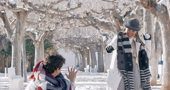 A cute cheerful couple in a winter park: the lady throwing a snowball on the guy