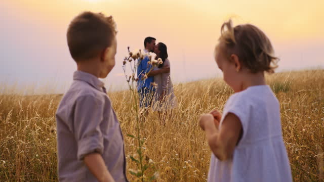 Slow motion footage of young kids playing with a flower in a golden meadow, parents hugging in the background