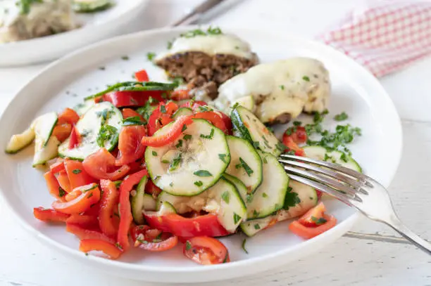 Mediterranean salad with zucchini, tomatoes and paprika. Marinated with olive oil, vinegar, honey and herbs. Served for dinner or lunch on a plate on white table background. Closeup and front view