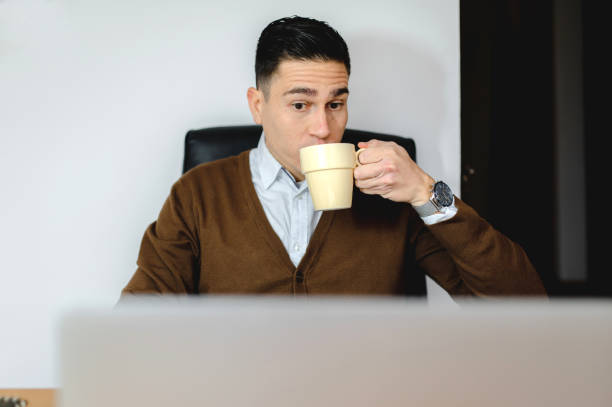istock Young man drinking from a mug looking at his laptop in the office 1459917979