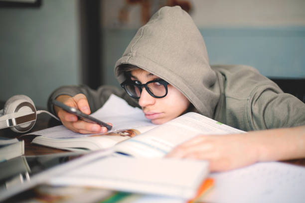 Tired boy with eyeglasses cheating and looking at his smartphone instead of doing homework A tired boy with eyeglasses cheating and looking at his smartphone instead of doing homework one teenage boy only stock pictures, royalty-free photos & images