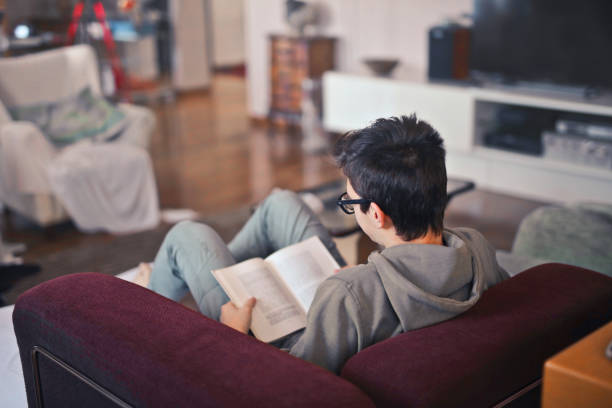 istock Young student with a grey outfit sitting on a red couch and reading 1459918549