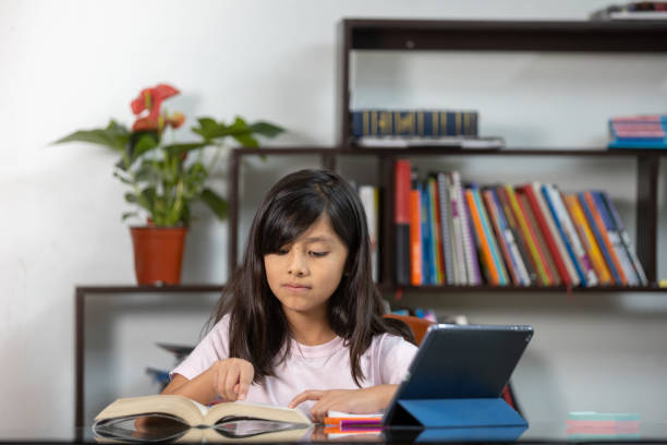 istock Hispanic schoolgirl reading a book and using a notebook and tablet at home - online education 1459918956