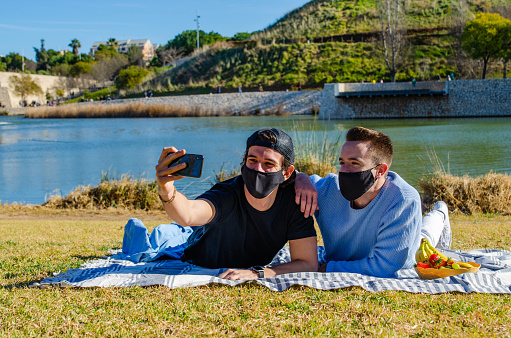 Happy Gay Couple with Protective Black Face Mask Taking Selfie with Smarphone Lying on Tablecloth Doing Picnic at the Park With Lake Behind