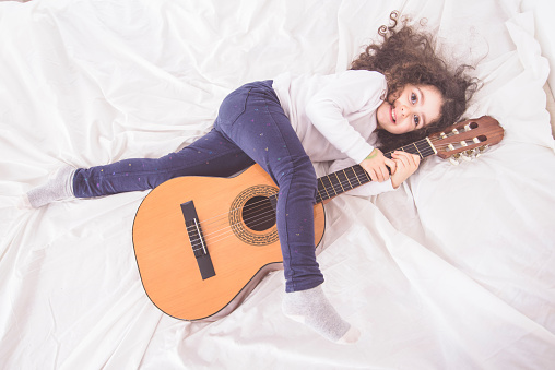 A top view of a cute Caucasian little girl lying on a bed with a guitar near her