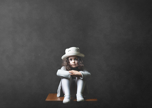 A closeup shot of a cute Caucasian little girl in a hat sitting on a chair and looking at the camera