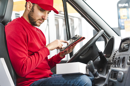 A delivery man with a red hat and sweater registering the details of the product sitting on the driver's seat in a vehicle