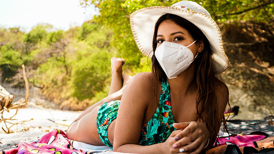 An attractive Latina woman lying on a sandy beach while wearing a face mask for covid-19 prevention