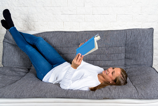 Young woman lying on a sofa smiling with her legs up and reading a book.