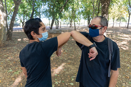 Stock photo of two male friends wearing face mask greeting each other with elbow bump in the park.