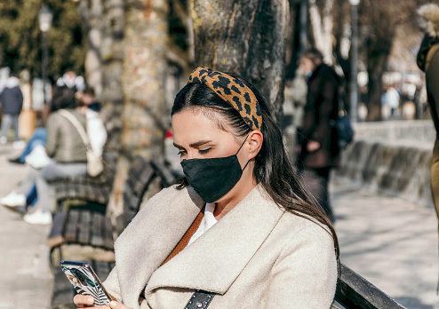 Young woman sitting on bench in city. Wearing mask, face mask, protective mask. Epidemic, corona virus, covid, using mobile phone, texting.